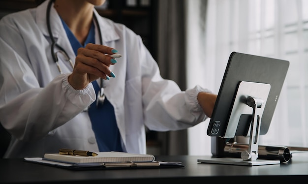 Serious female doctor using laptop and writing notes in medical journal sitting at desk Young woman professional medic physician wearing white coat and stethoscope working on computer at workplace