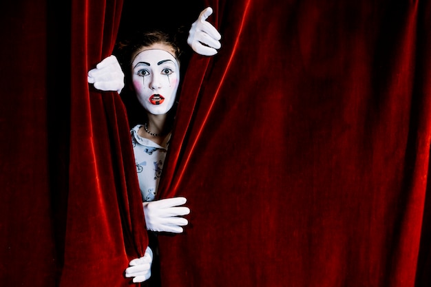 Serious female mime artist peeking from red curtain