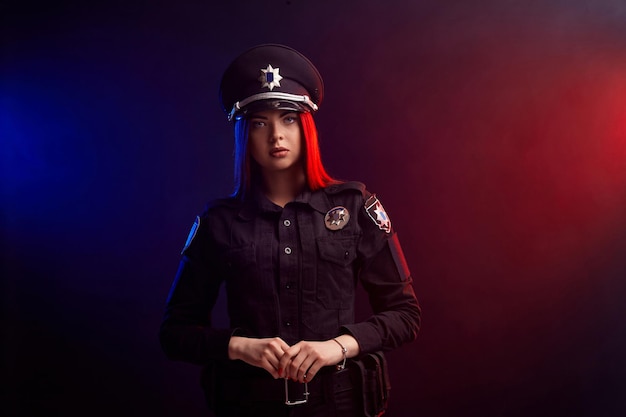 Serious female police officer is posing for the camera against a black background with red and blue ...