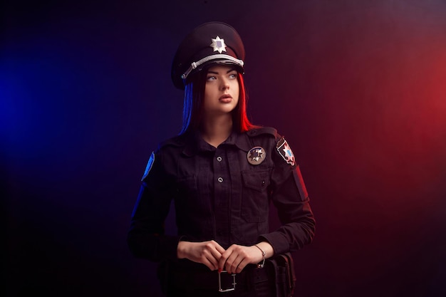 Serious female police officer is posing for the camera against a black background with red and blue ...