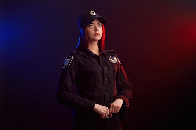 Serious female police officer is posing for the camera against a black background with red and blue ...