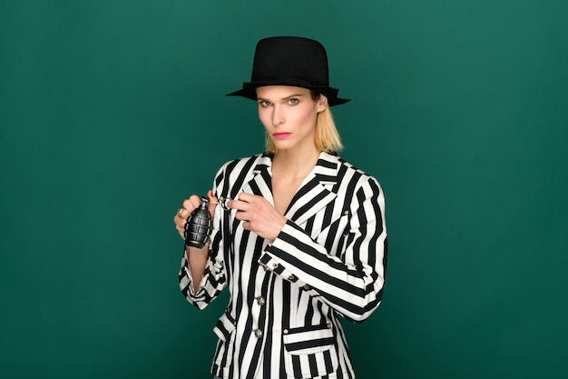 Serious female in stylish striped jacket and hat looking at camera and pulling grenade pin against green background