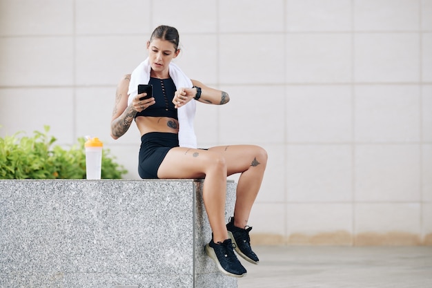 Serious fit woman resting after training and checking her heart rate and overall activity via applications on smartphone and smartwatch