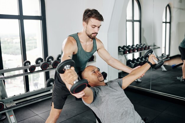 Serious fitness trainer helping mature man to do dumbbell fly on bench in gym