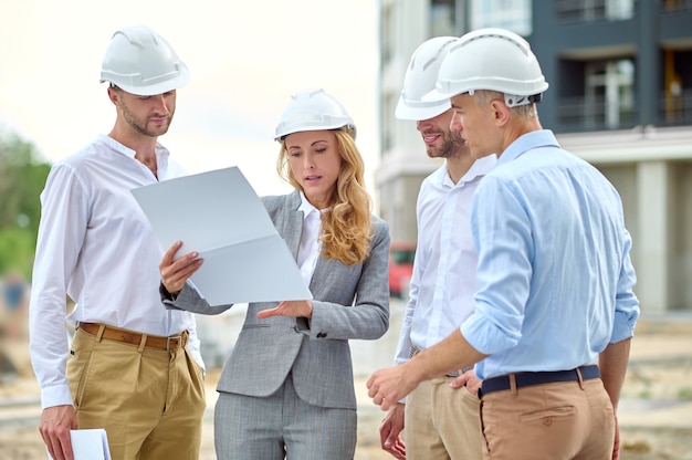 Supervisore femminile biondo serio e concentrato che controlla i documenti di costruzione circondato da costruttori maschi in caschi protettivi