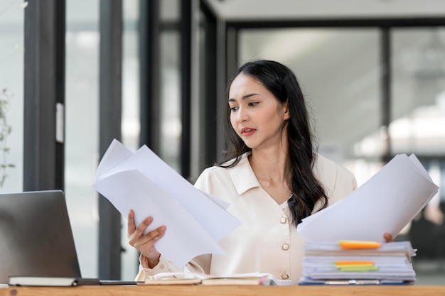 Serious focused businesswoman holding papers preparing report analyzing work results female executive doing paperwork at workplace using computer looking busy for data analysis