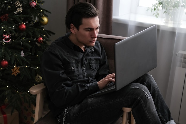 Serious focused busy young man is using a laptop while sitting in an armchair