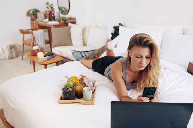 Serious frowning beautiful young woman resting on bed, eating breakfast, watching show on laptop and texting friend or checking social media