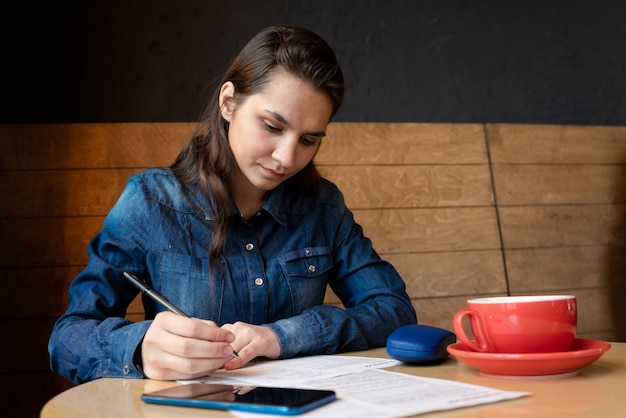 Foto modello di ragazza seria firma il rilascio, c'è una tazza rossa sul tavolo, custodia per occhiali blu e uno smartphone, una donna con una camicia di jeans