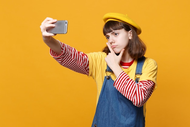프랑스 베레모를 입은 진지한 10대 소녀, 휴대폰으로 셀카를 찍는 데님 선드레스, 노란색 배경에 격리된 턱에 손을 올려놓습니다. 사람들이 감정, 라이프 스타일 개념입니다. 복사 공간을 비웃습니다.