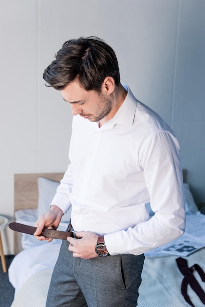 Serious goodlooking man dressing while standing near bedding at home