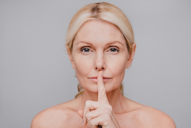 Serious goodlooking mature woman with clear skin making silent\
gesture isolated over grey