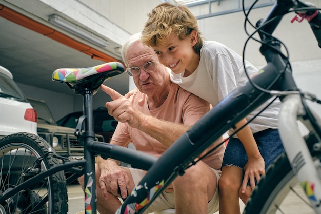 진지한 할아버지가 소년에게 자전거 수리 방법을 설명하고 주차장에서 활짝 웃으며 주의 깊게 듣고 있다
