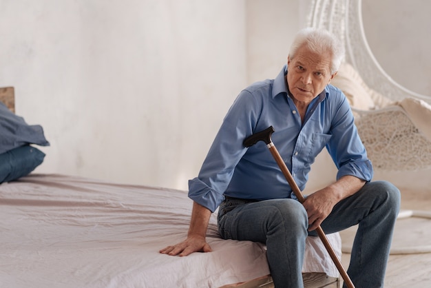 Serious grey haired elderly man sitting alone in the room and holding a walking stick while looking at you