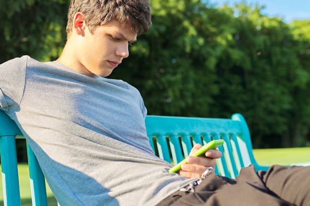 Serious guy teenager using smartphone having rest sitting on bench in park