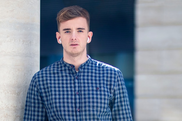 Serious guy, young handsome man in shirt listening to music in wireless earphones or headphones,  outdoors. Hipster boy with modern gadget, copy space