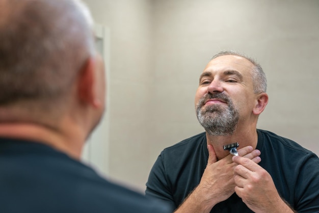 Photo serious handsome man looking in mirror and shaving his whiskers