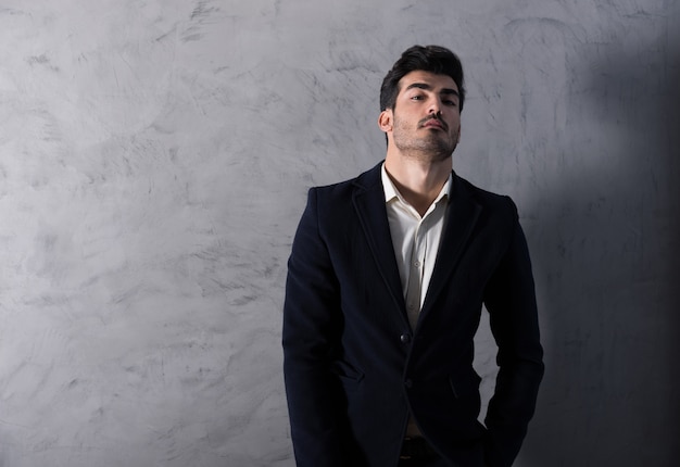 A serious handsome young businessman in a black suit standing in front of a grey wall