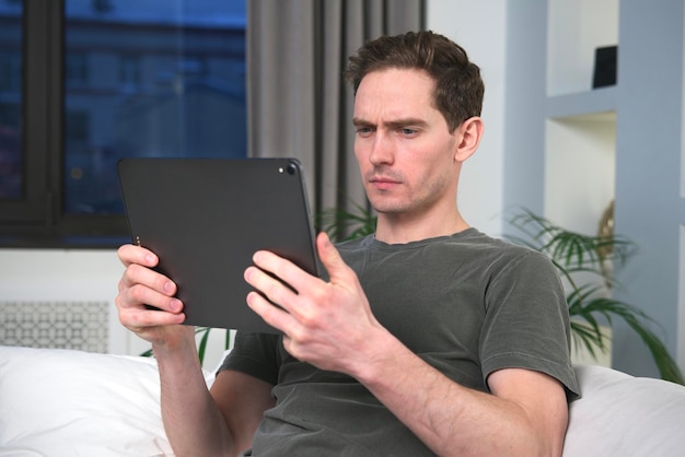 Serious handsome young man is using tablet computer gadget at home reading or watching movie sitting at home in living room on couch