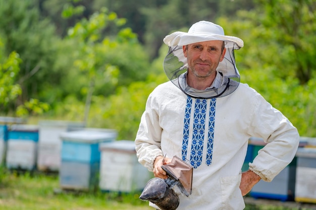 Серьезный трудолюбивый пчеловод позирует перед камерой на пасеке. Человек с курильщиком пчелы в руках. Концепция пчеловодства.