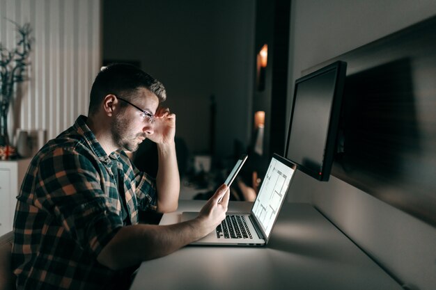 Impiegato non rasato laborioso serio con gli occhiali facendo uso della compressa mentre sedendosi a tarda notte nell'ufficio