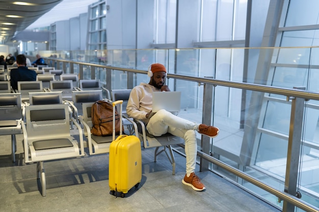 Serious hipster black man sitting in airport terminal using laptop wear headphones listening music