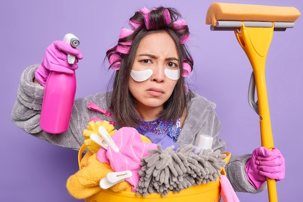 Serious housewife focused at camera applies beauty patches\
under eyes to reduce wrinkles makes hairstyle holds cleaning mop\
and bottle of detergent dressed in home clothes poses near laundry\
basket