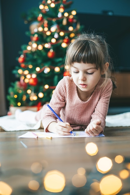 Serious little girl 56 years old lying by the holiday tree putting her hopes and dreams into a letter for Santa Claus