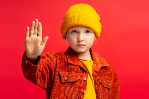 Photo serious little girl wearing yellow hat showing stop gesture with hand isolated on red background