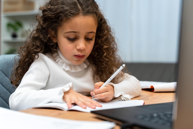 Serious little schoolgirl sitting at table and preparing her home tasks while doing homework with the laptop Children education and distant learning concept Stock photo