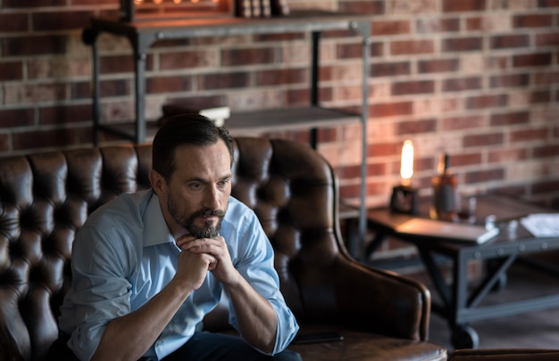 Serious look. Attractive thoughtful stylish man holding his chin and looking thoughtfully while sitting on the couch