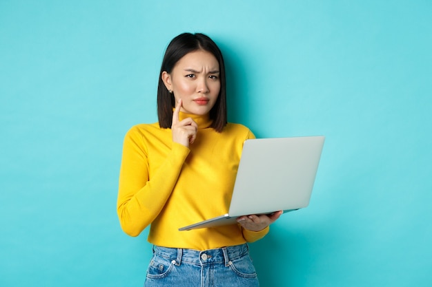 Donna asiatica dall'aspetto serio che lavora al computer portatile e pensa, aggrottando le sopracciglia alla telecamera, risolvendo problemi sul lavoro, in piedi su sfondo blu.