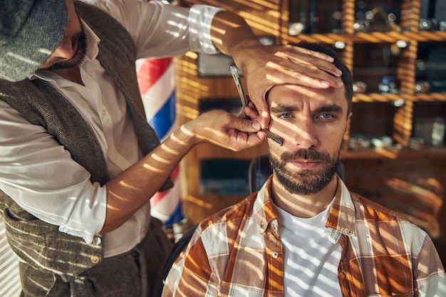 Serious-looking young man getting shaved with a straight razor by a professional barber