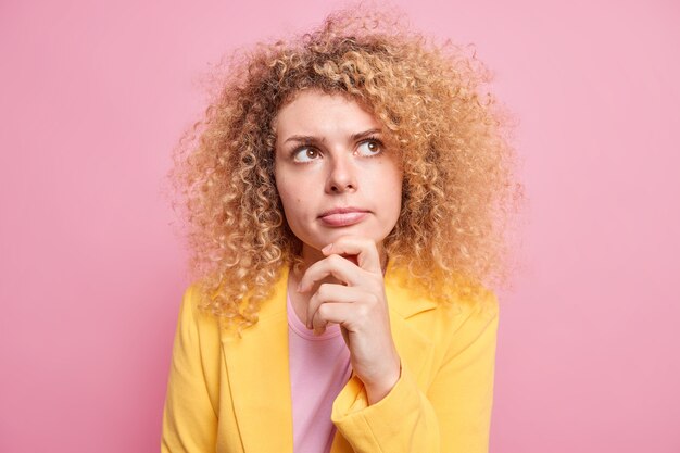 Serious looking young woman with curly hair holds chin looks above thinks deeply focused on something ponders suspicion situation dressed in formal yellow jacket isolated over pink wall