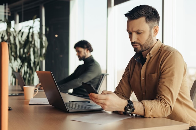 Serious male company employee checking mobile phone using laptop working with male colleague at open