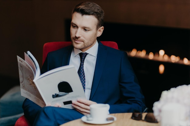 Serious male entrepreneur reads business magazine sits in\
coffee shop drinks coffee or tea dressed in formal suit has dinner\
break and rest after being in office business and lifestyle\
concept