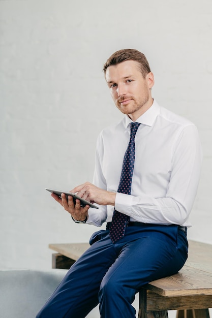 Serious male entrepreneur watches video on digital tablet\
messages with business partners dressed elegantly sits on wooden\
table poses against white wall businessman updates banking\
account