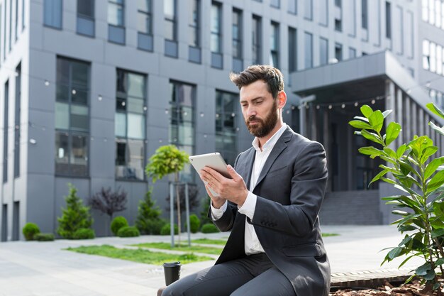 Libero professionista maschio serio in giacca e cravatta che guarda un corso online usa un tablet, seduto in un luogo pubblico su una panchina