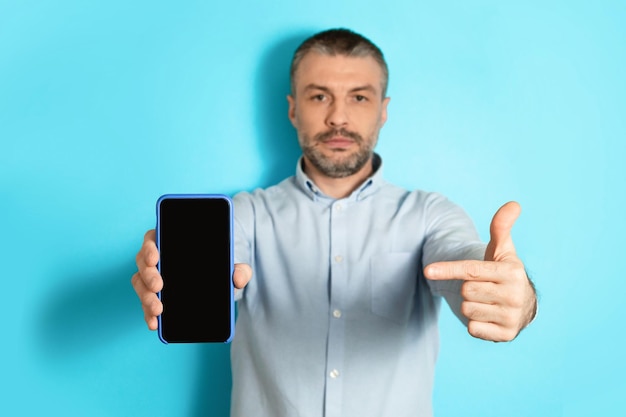 Serious male showing cellphone screen pointing finger over blue background