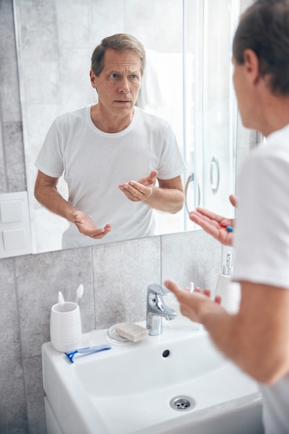 Photo serious male with a pair of capsules in his hand staring in the bathroom mirror