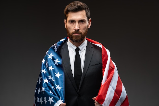 Serious man of adult age with pleasant appearance wrapped in\
usa flag and looking at the camera attentively. politics debate\
concept