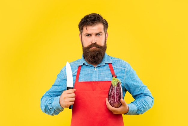 Serious man in apron holding chefs knife and eggplant yellow background chef