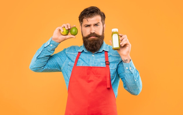 serious man in apron holding limes and juice bottle yellow background juice shopkeeper