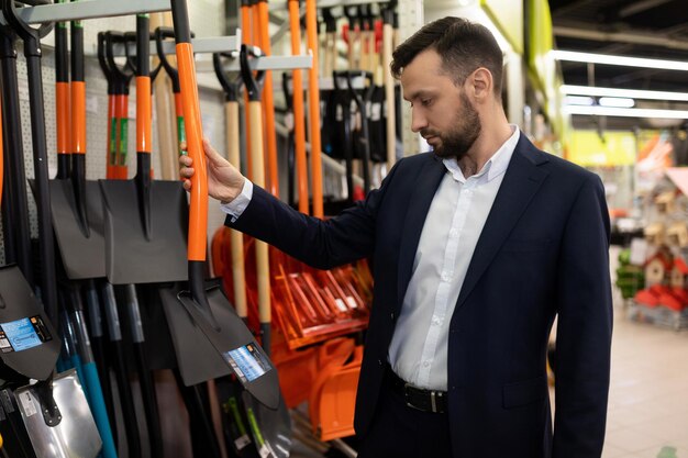 A serious man in a business suit chooses a shovel in a gardening store