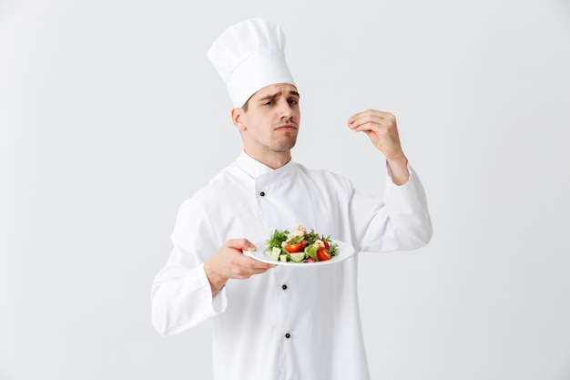白い壁に隔離されたプレートに新鮮なグリーンサラダを示す制服を着た真面目な男のシェフ料理人