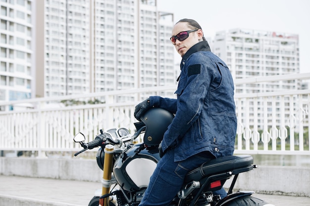 Serious man in denim jacket and sunglasses sitting on motorcycle and turning back to camera