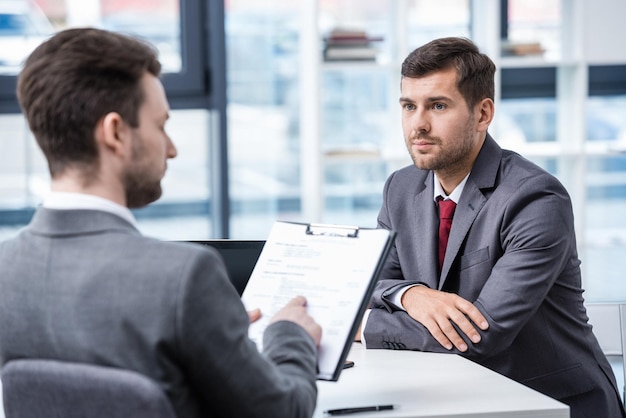 Serious man in formal wear looking at businessman with clipboard during job interview, business