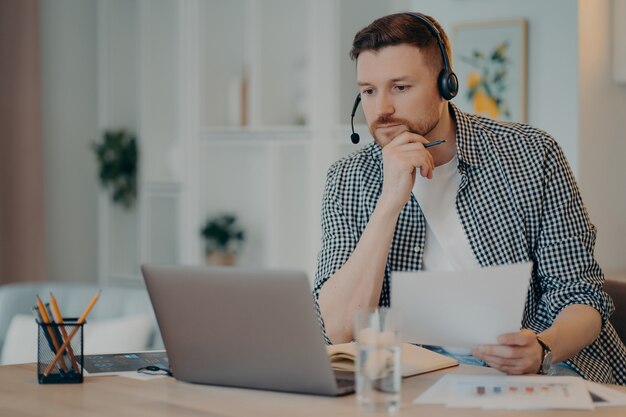 Serious man freelancer in casual wear holding document in hand and looking at laptop screen, analyzing information while working online in living room. Remote work and freelance concept