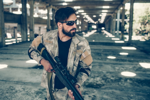 Serious man in glasses is standing in hangar and holding paintball gun in hands.