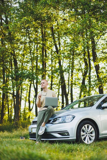 A serious man in glasses is working on a laptop on the hood of his car Remote work in nature Work on a laptop online Working on a computer while traveling